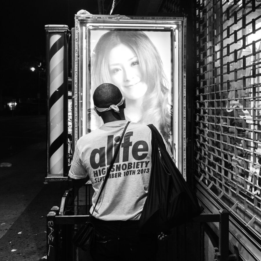 Black-and-white frame of a man in an Alife t-shirt standing before a glowing barbershop poster of a smiling woman