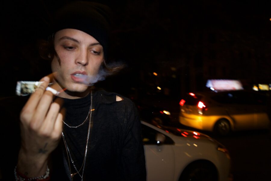 Young man in a black beanie exhaling cigarette smoke at night, blurred traffic lights behind him