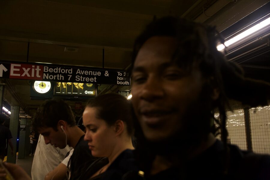 Subway platform at Bedford Avenue, a face passing close to the lens out of focus, exit sign overhead