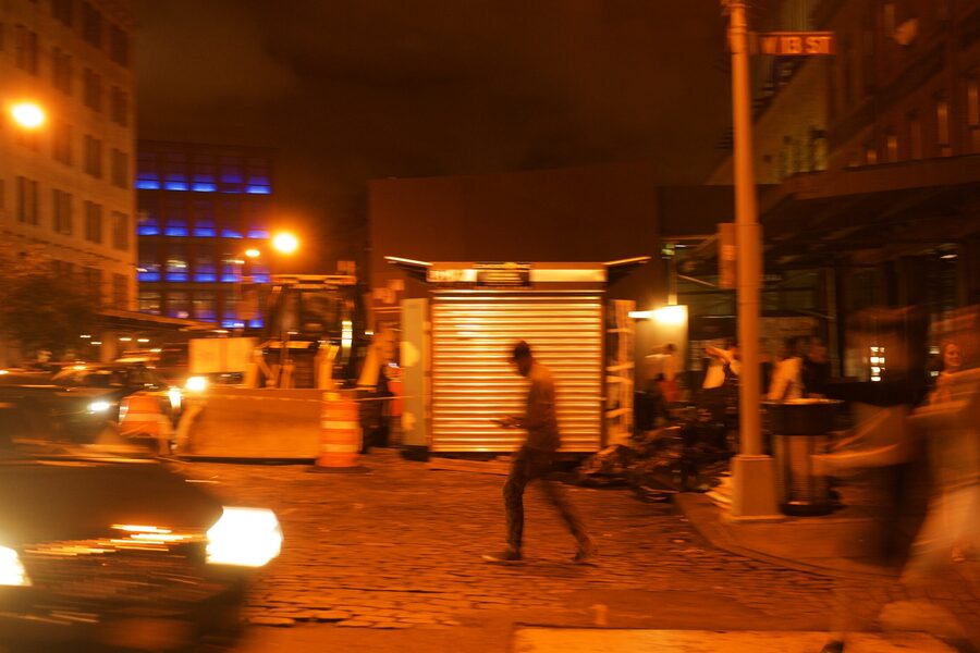 Figure walking past a shuttered kiosk on West 13th Street, everything washed in orange sodium light