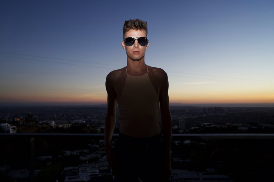 Man in aviator sunglasses standing above the Los Angeles basin at dusk, sky fading from orange to blue