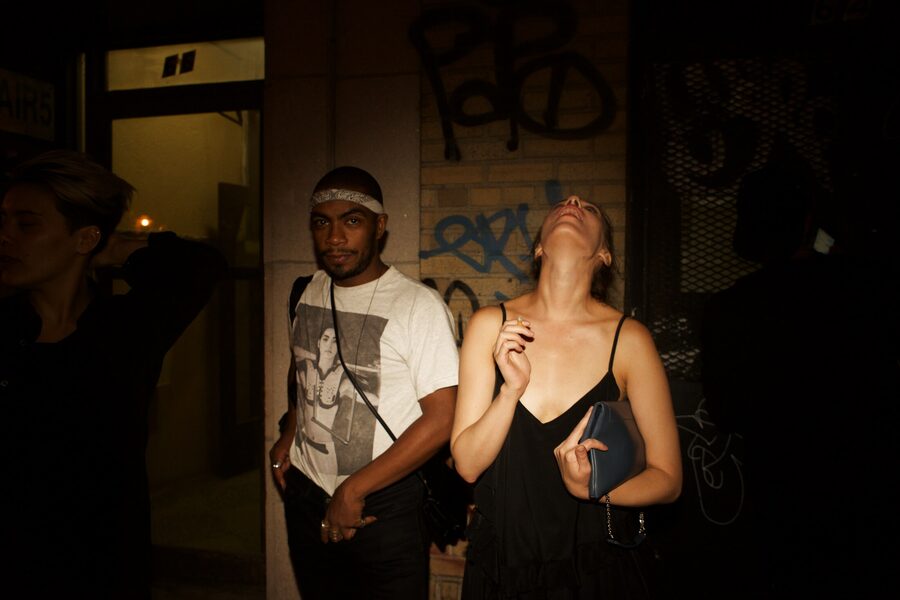 Direct-flash frame of a woman in a black slip dress throwing her head back beside a man in a graphic tee, graffiti behind them