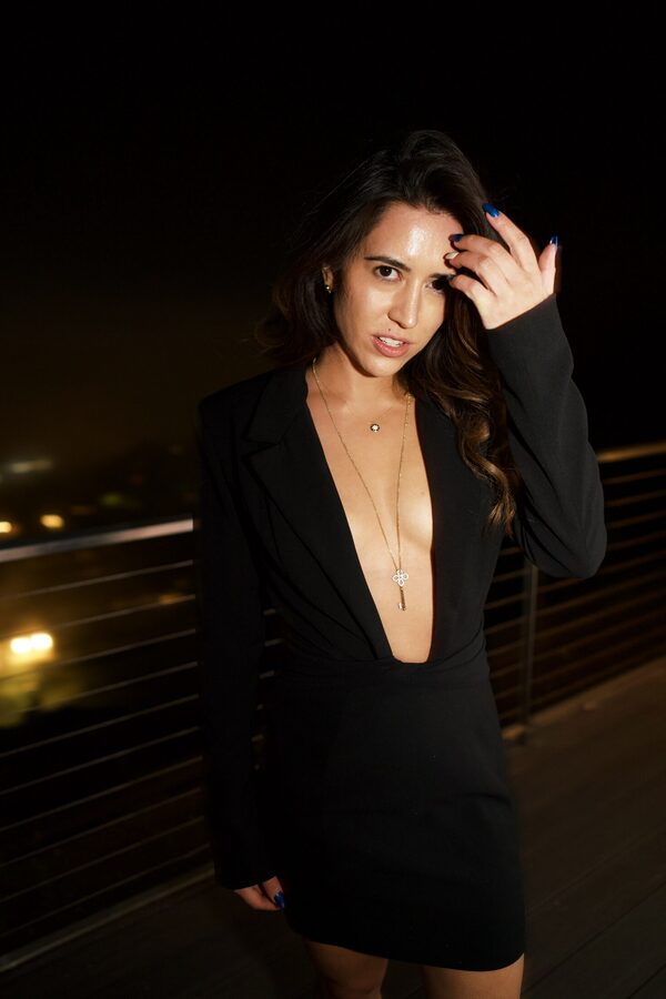 Woman in a deep-cut black blazer on a rooftop at night, a single streetlight burning far behind