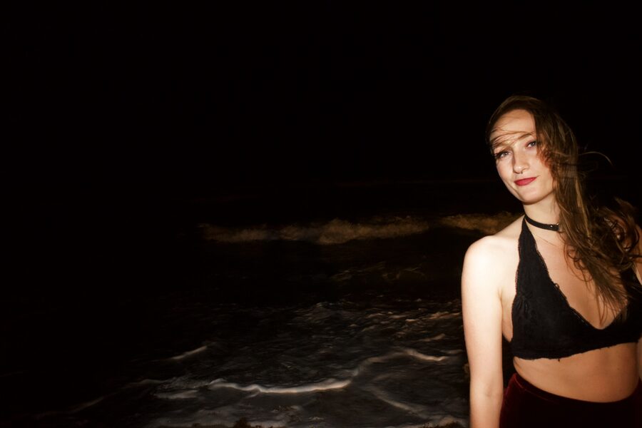 Woman standing in dark ocean surf at night, lit by direct flash, waves breaking white behind her