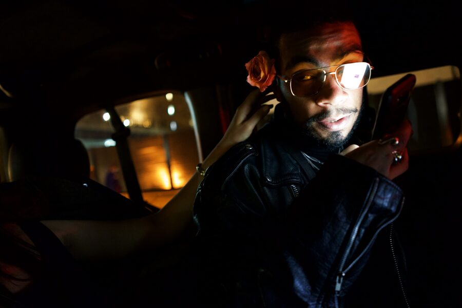 Man in a leather jacket and tinted glasses, a rose tucked behind his ear, face lit by his phone in the back of a car