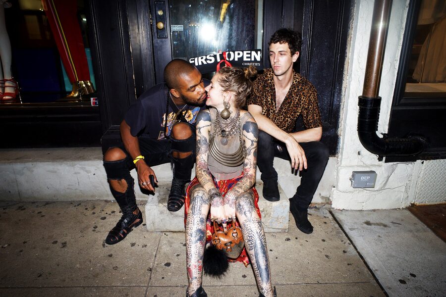Three friends collapsed on a storefront stoop at night, a heavily tattooed woman leaning back between them