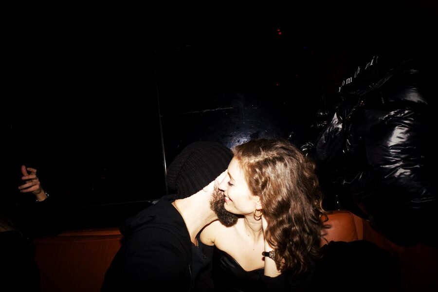 Couple kissing in a dark banquette, his beanie and beard against her cheek, flash catching a puffer jacket behind them