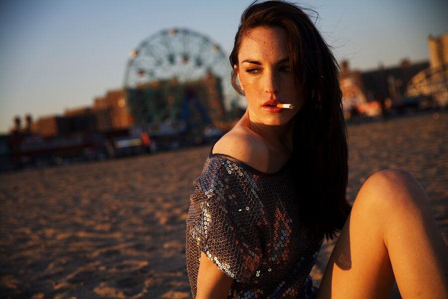 Woman in a sequin dress on the Coney Island sand at dusk, cigarette in her lips, the Wonder Wheel soft behind her